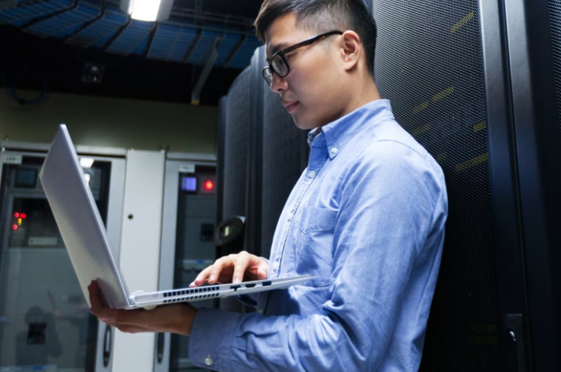 An IT professional is using a laptop in a server room.